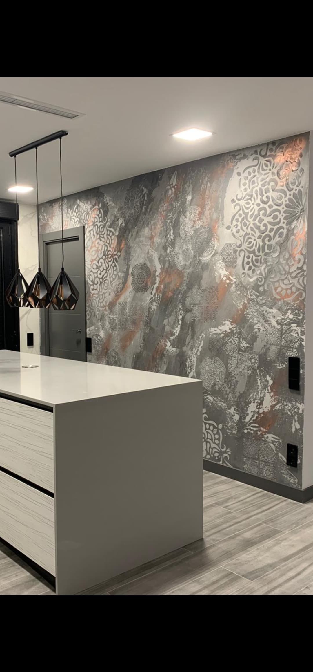 Mural decorativo en relieve con mandalas moriscas en gris, blanco y cobre cubriendo la pared de una cocina abierta, junto a una isla blanca y lámparas colgantes negras