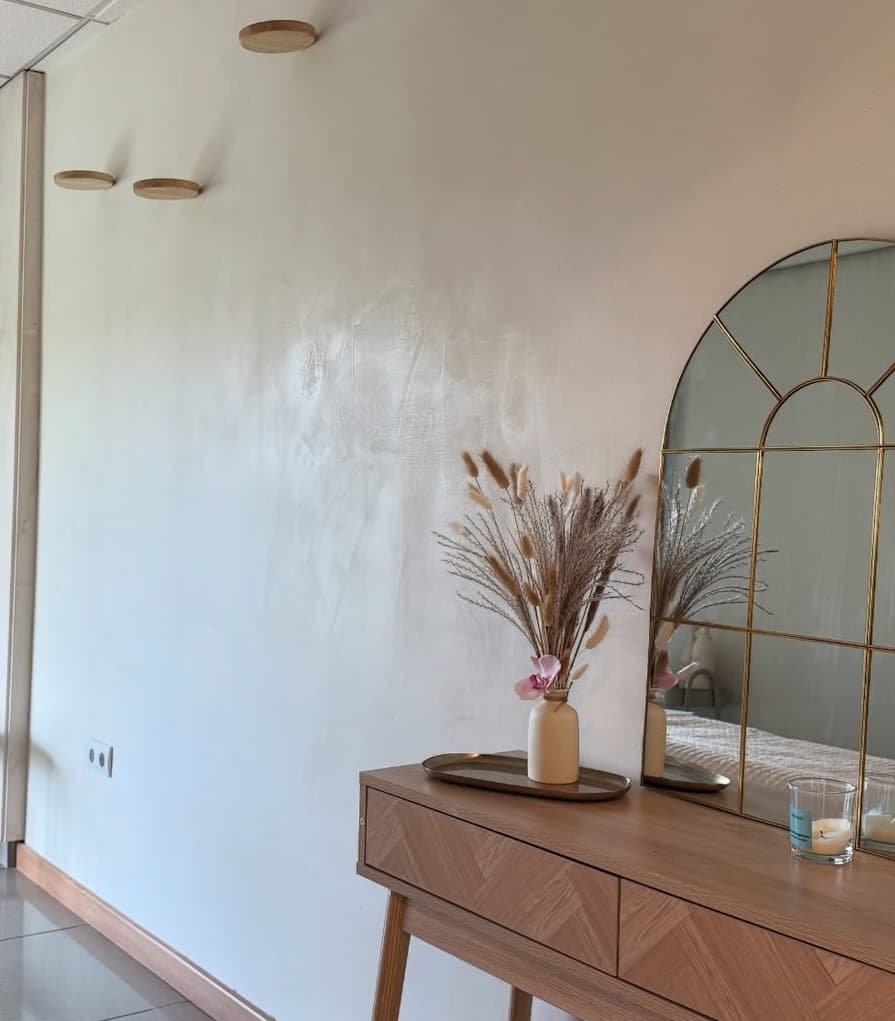 Pared de recibidor con acabado de estuco texturizado en blanco, consola de madera con espejo de arco y jarrón de flores secas
