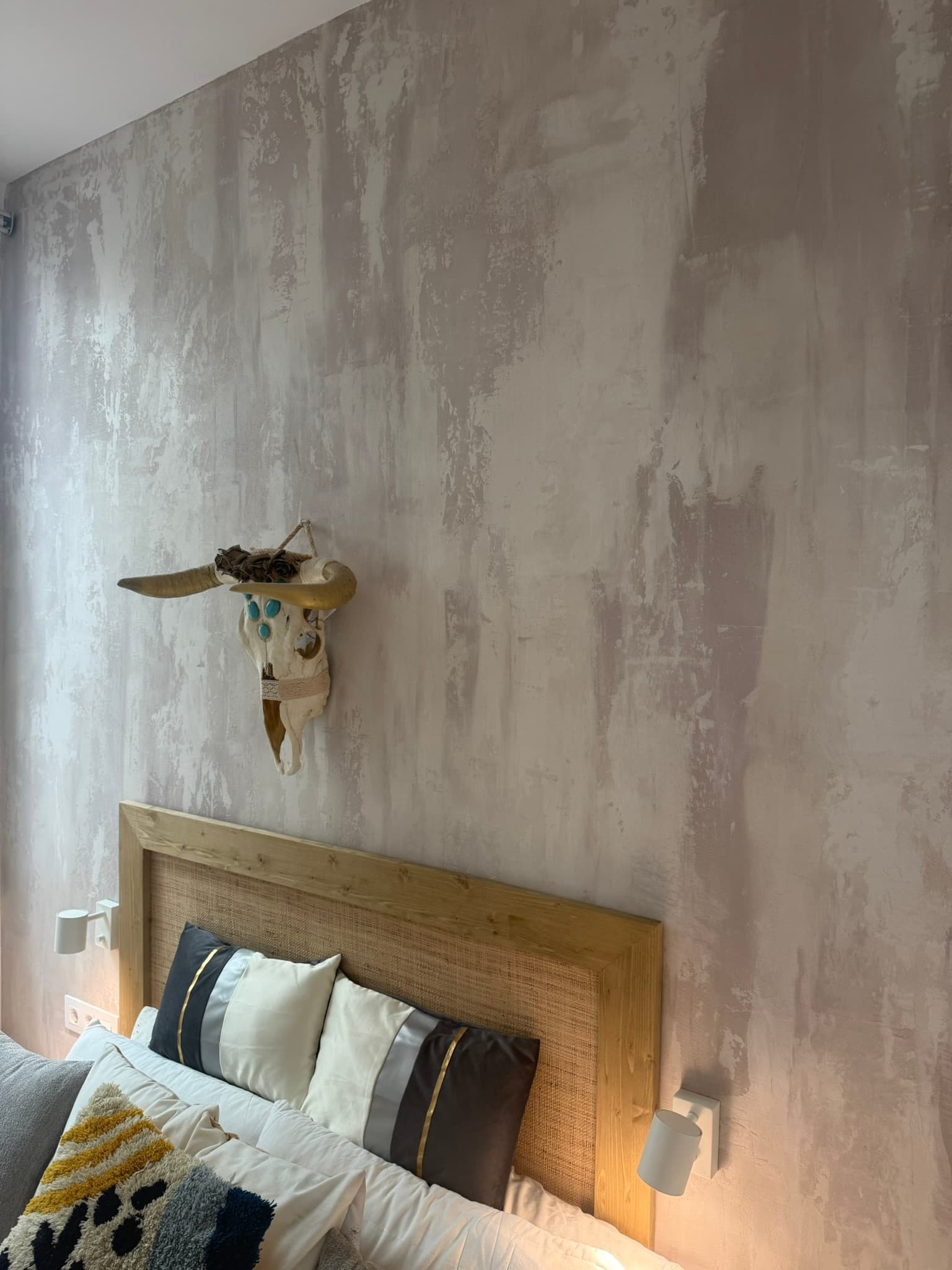 Detalle de pared en estuco gris veteado con calavera de vaca decorativa colgada, sobre un cabecero de madera y ratán