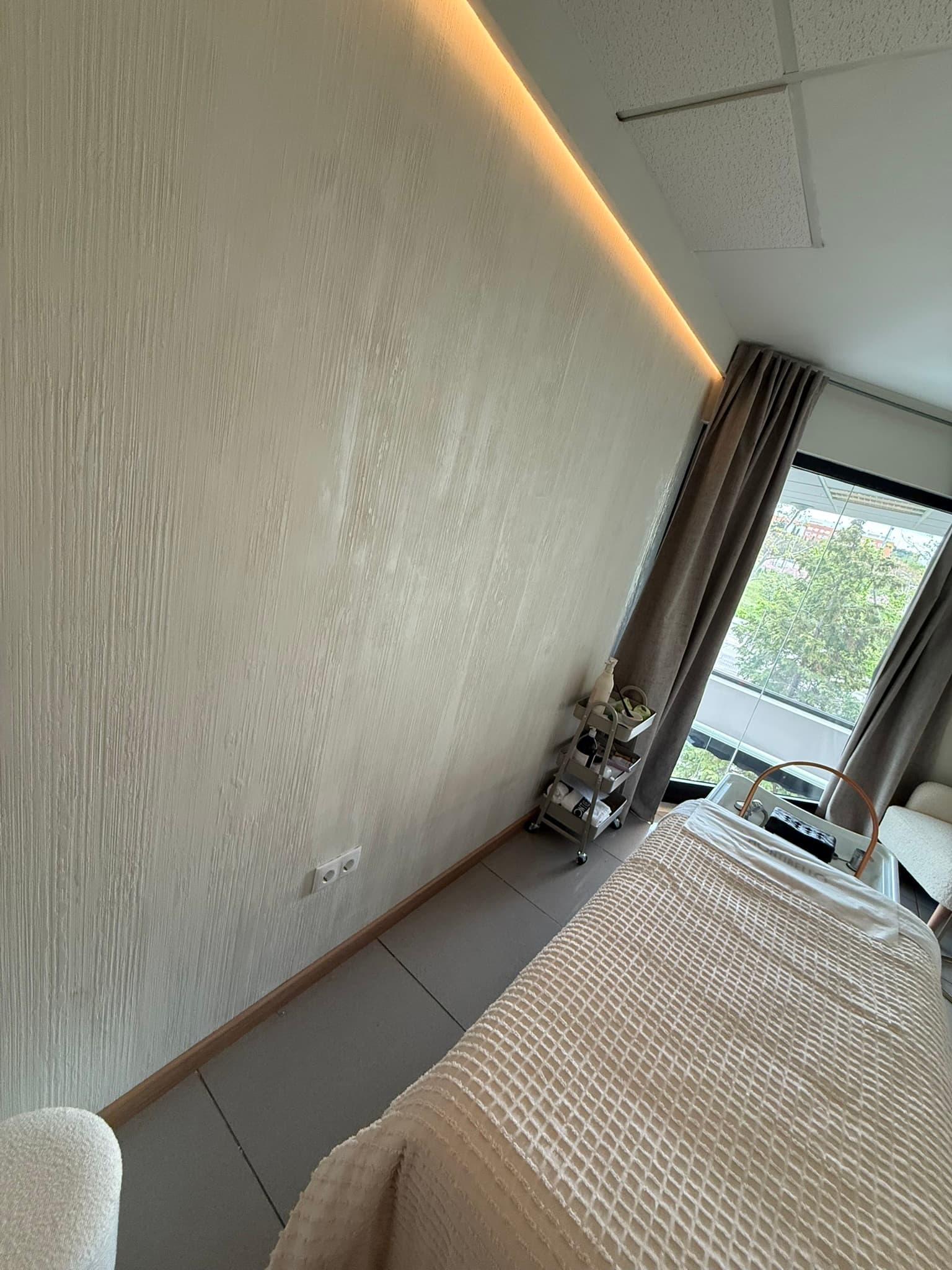 Sala de tratamientos con pared en estuco crema, carrito auxiliar y gran ventanal con cortinas hasta el suelo