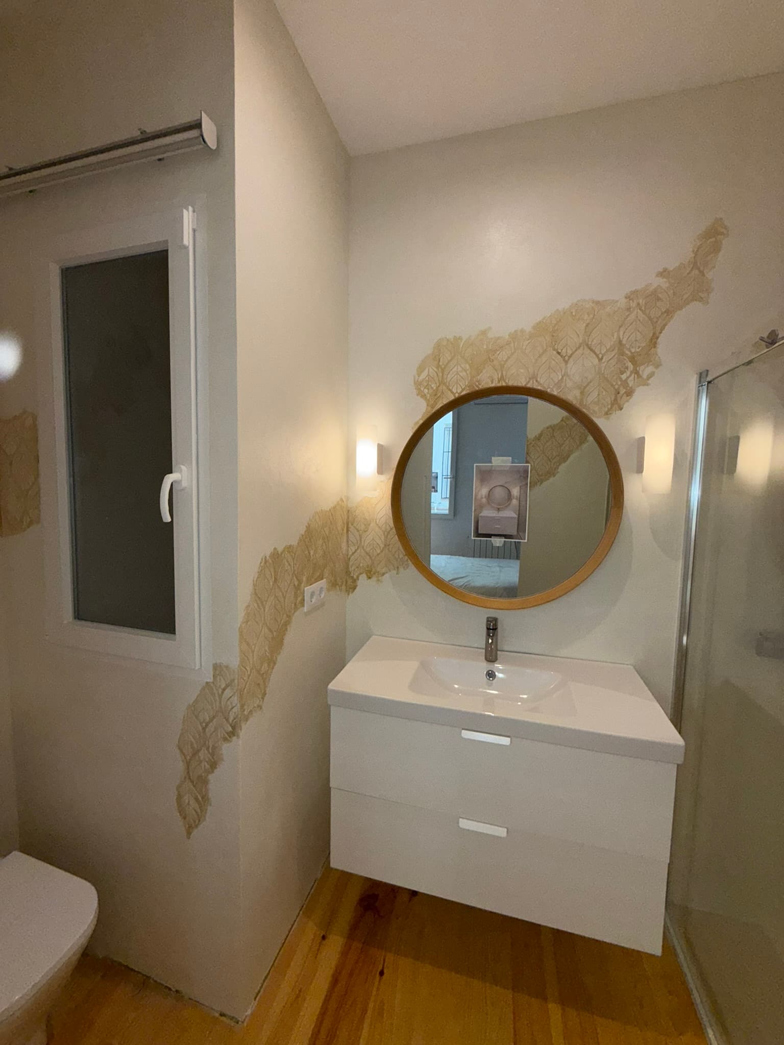 Baño con mueble de lavabo suspendido, espejo redondo y fragmentos de mural decorativo en relieve con hojas y pan de oro sobre la pared