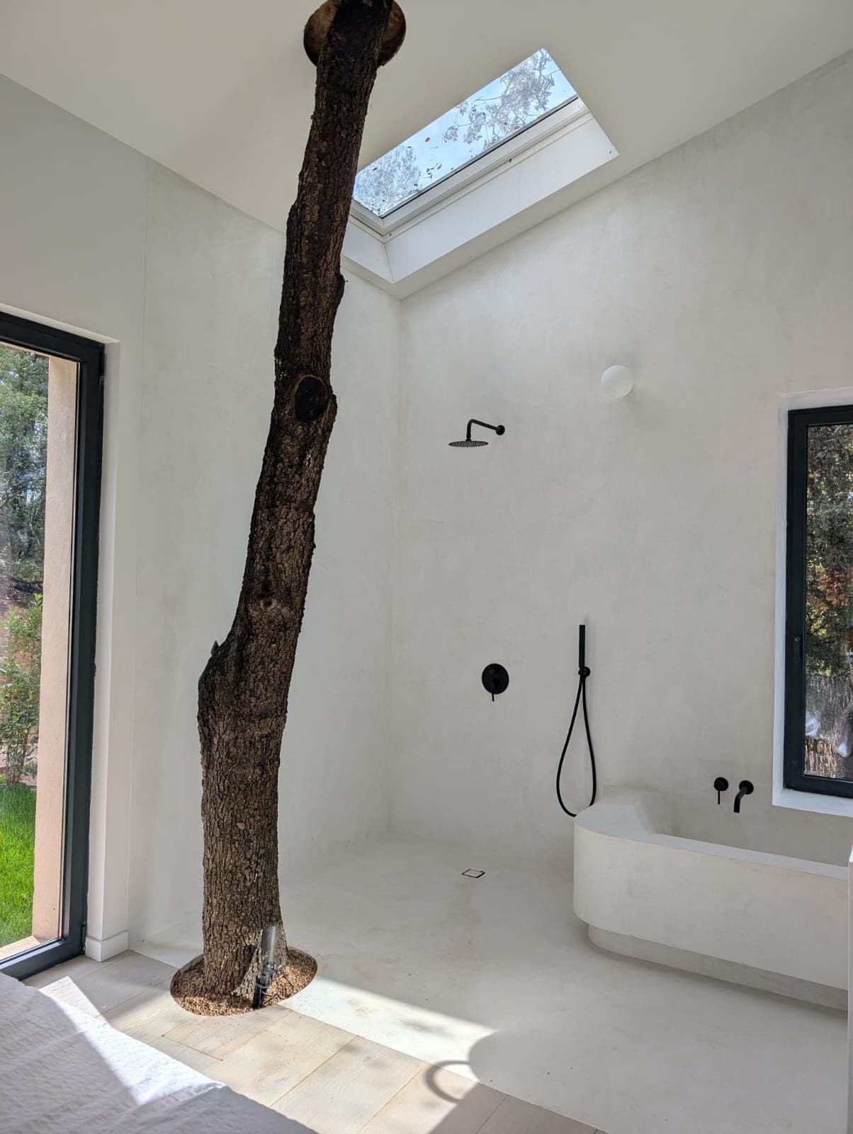 Ducha y bañera exenta en microcemento blanco sin juntas, con ducha de lluvia negra y ventanales de suelo a techo con vistas al jardín
