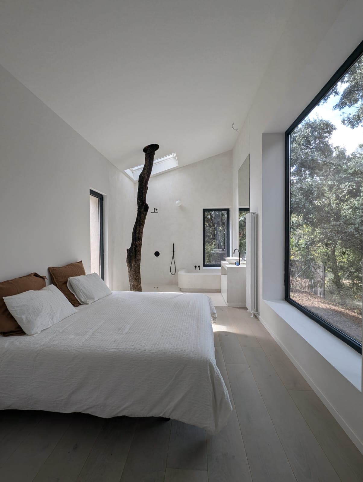 Dormitorio con suite de baño abierta en microcemento blanco continuo, bañera exenta y gran ventanal con vistas al bosque