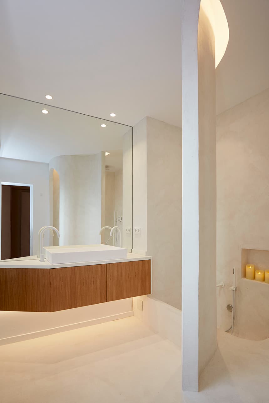 Lavabo de microcemento con encimera blanca y mueble de madera, junto a una pared curva en microcemento crema con hornacina portavelas