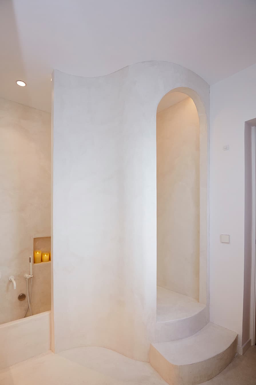 Entrada de ducha con arco escultórico en microcemento blanco de formas curvas, integrado en el muro del baño