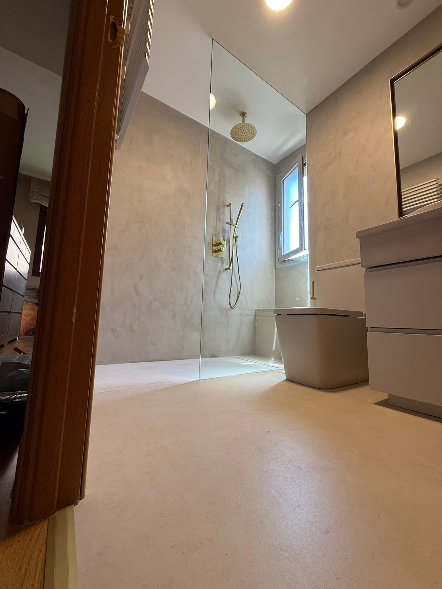 Ducha en microcemento gris con grifería dorada, ventana y suelo continuo sin juntas