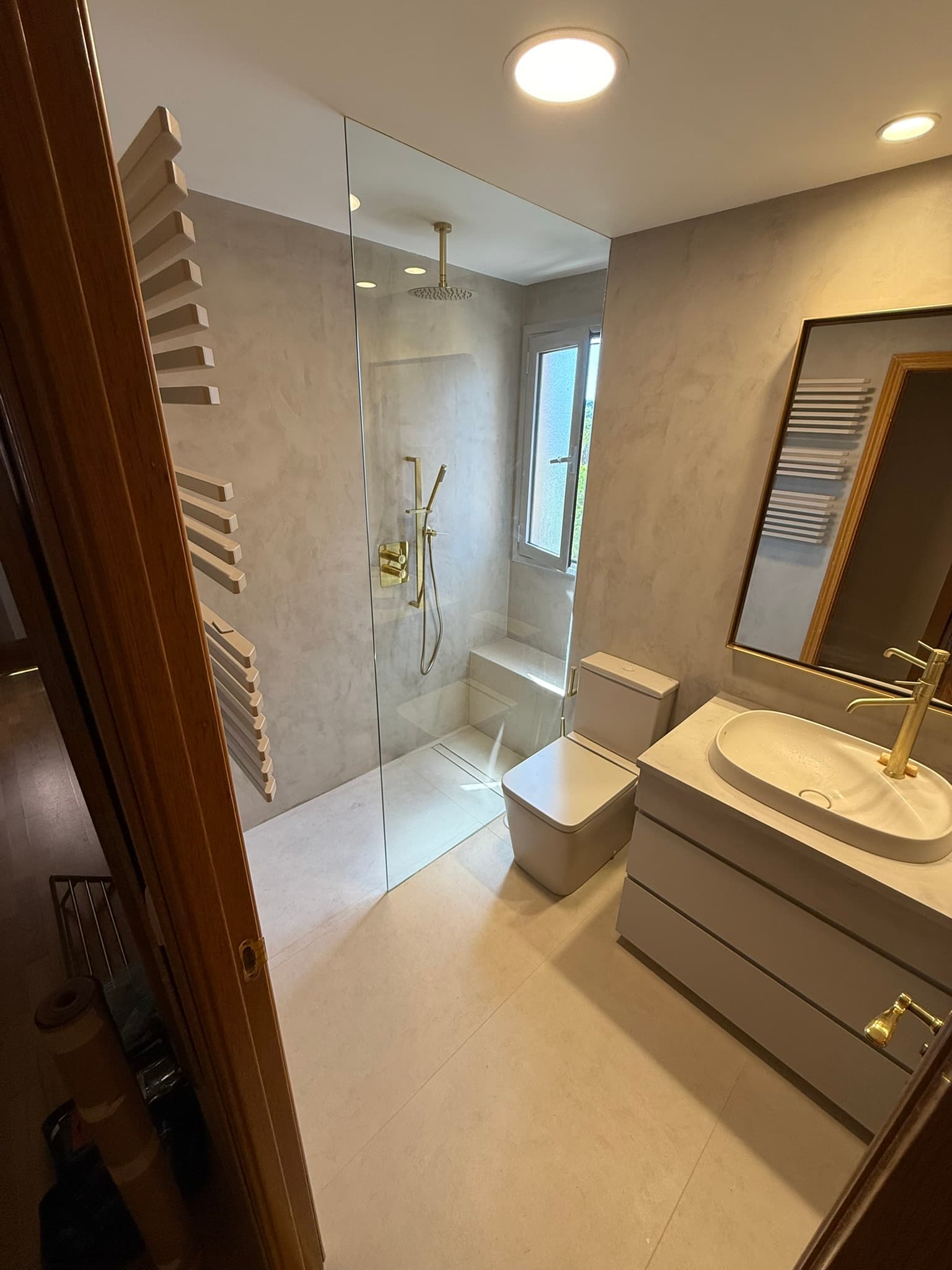 Baño en microcemento gris con ducha de obra y grifería dorada, junto a inodoro y mueble de lavabo