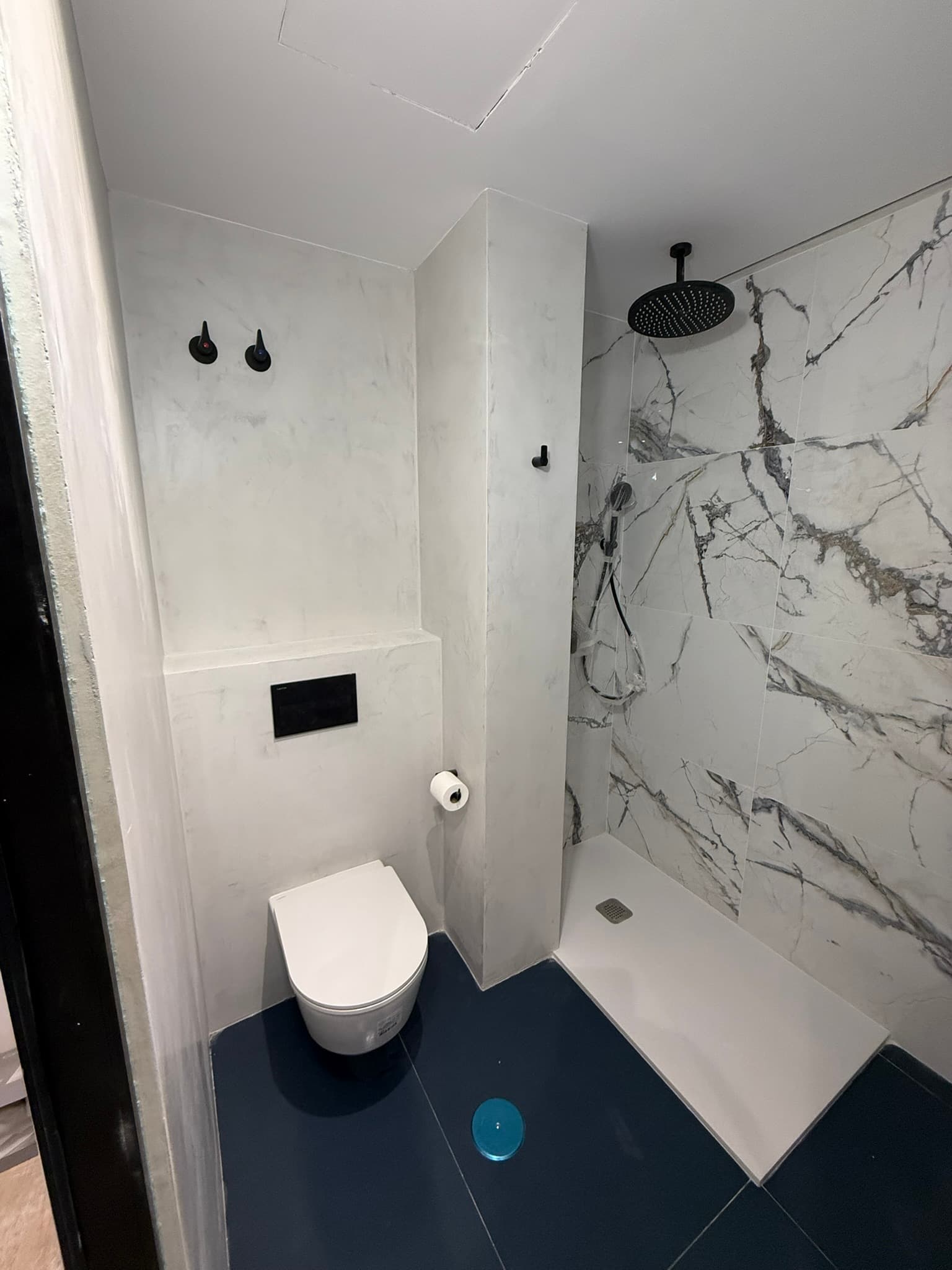 Baño combinando microcemento blanco con ducha alicatada en mármol, inodoro suspendido negro y suelo en azul marino
