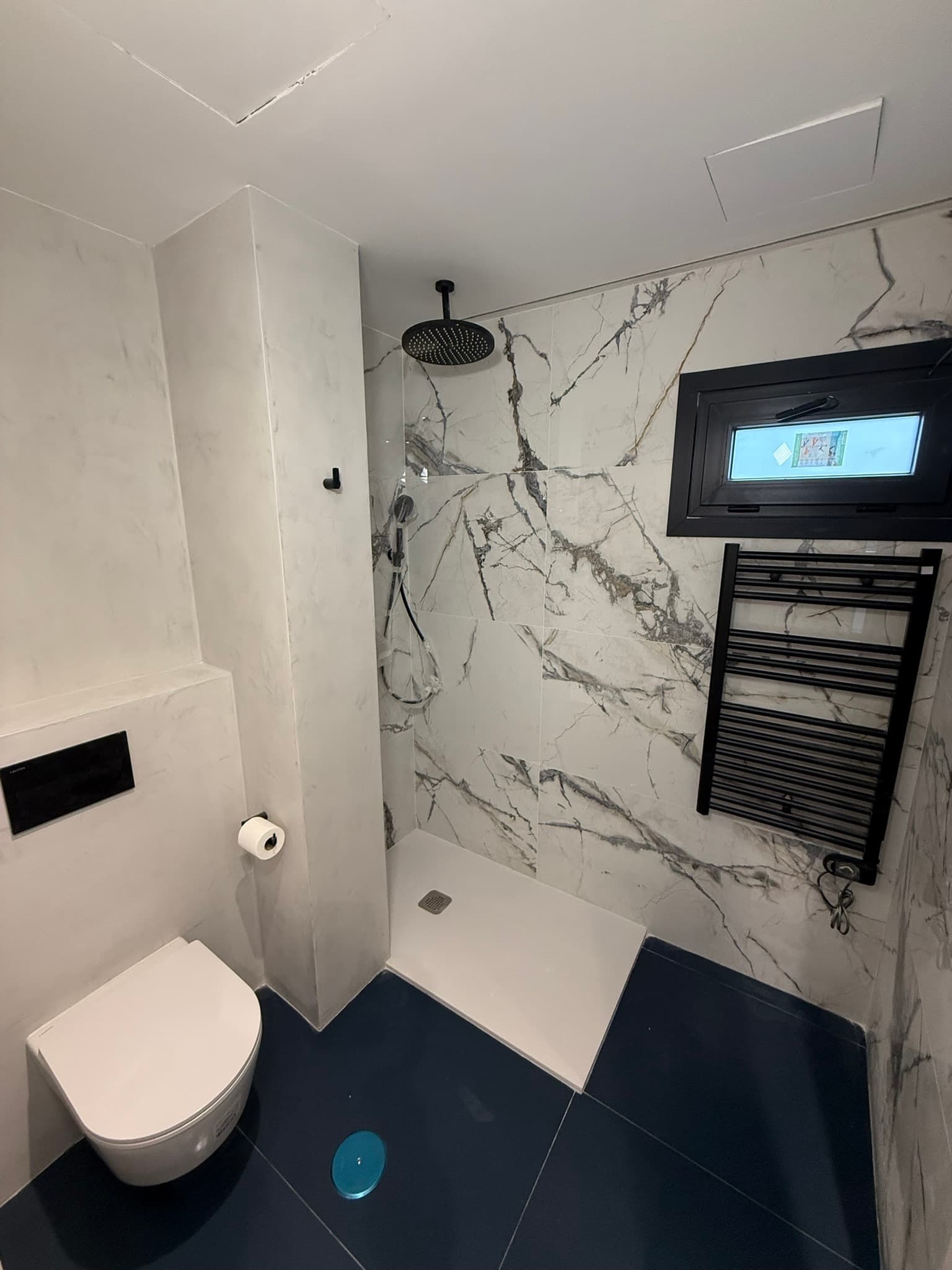 Ducha con paredes de mármol veteado y radiador toallero negro, combinada con microcemento blanco en el resto del baño