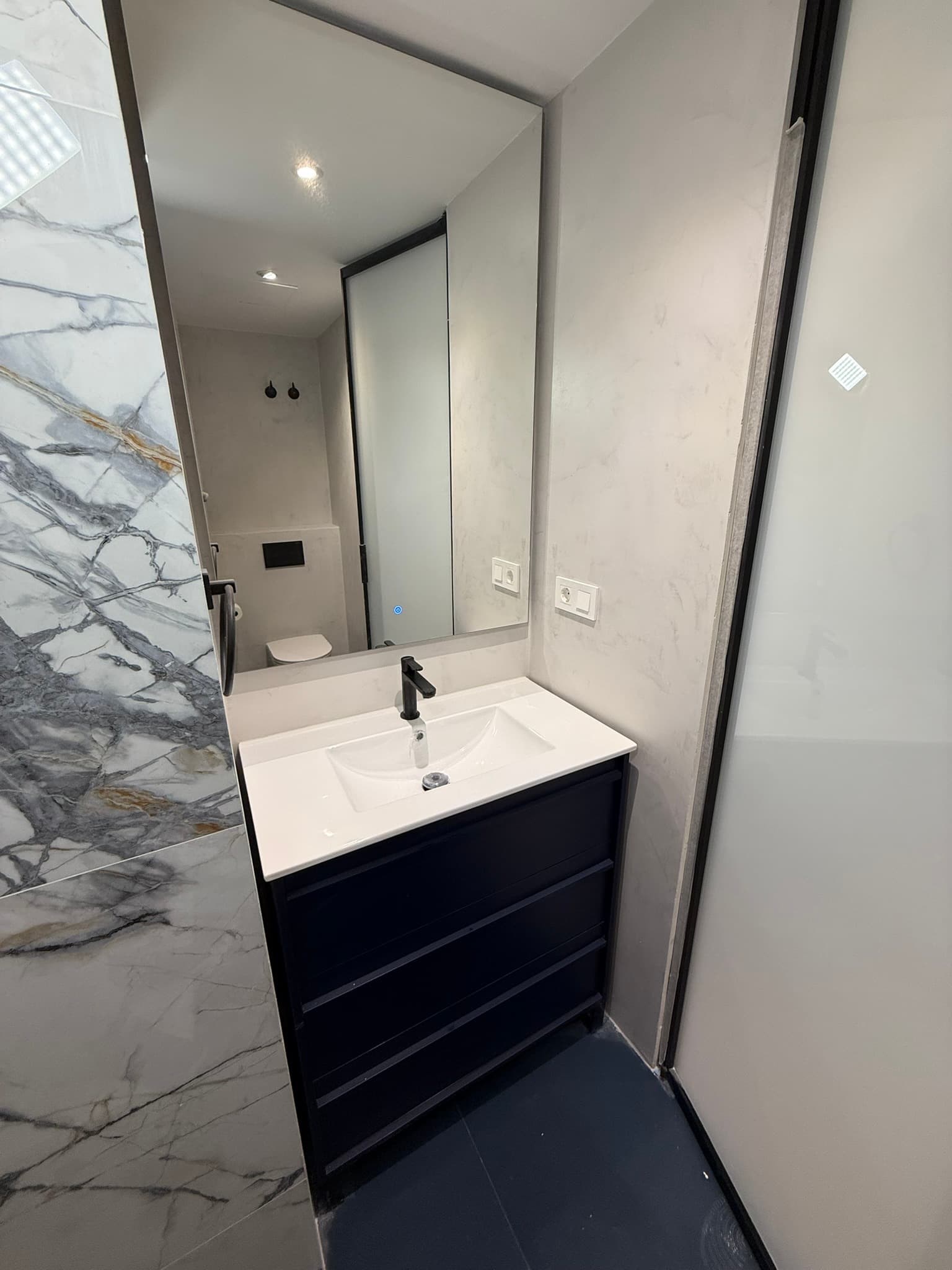 Lavabo de microcemento con mueble negro y encimera de madera, reflejando en el espejo la mampara de ducha con paredes de mármol