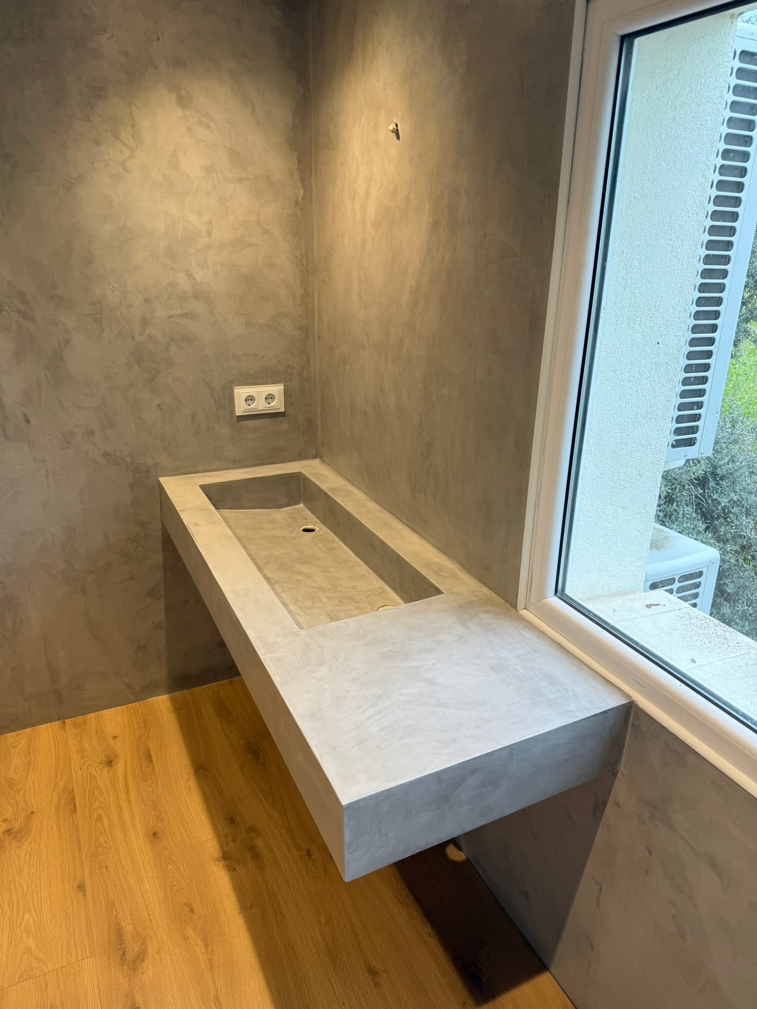 Lavabo corrido en microcemento gris con doble desagüe, pared continua sin juntas y ventana con vistas a la vegetación