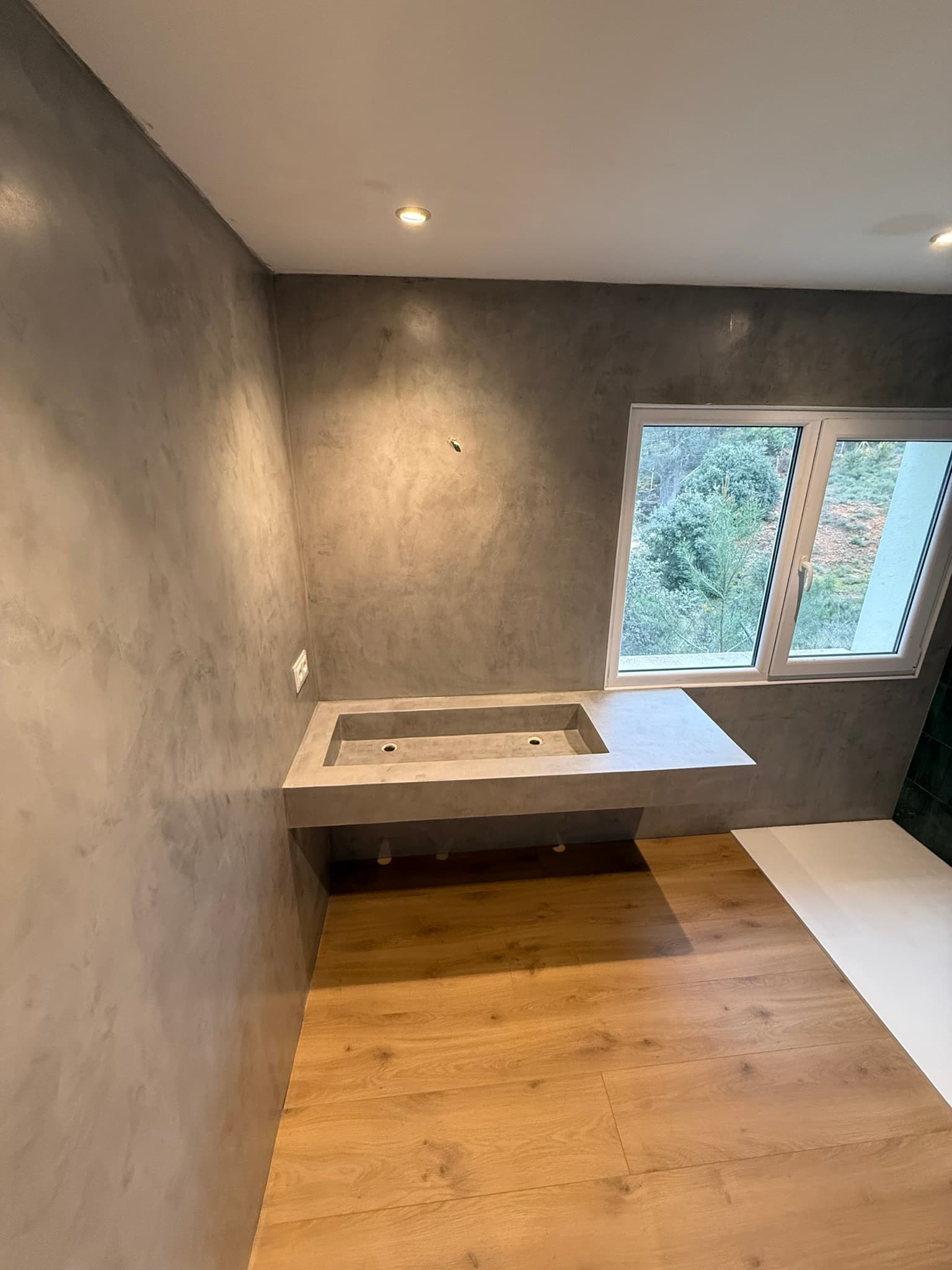 Detalle de lavabo corrido en microcemento gris junto a una ventana, con suelo de madera en la zona del dormitorio contiguo