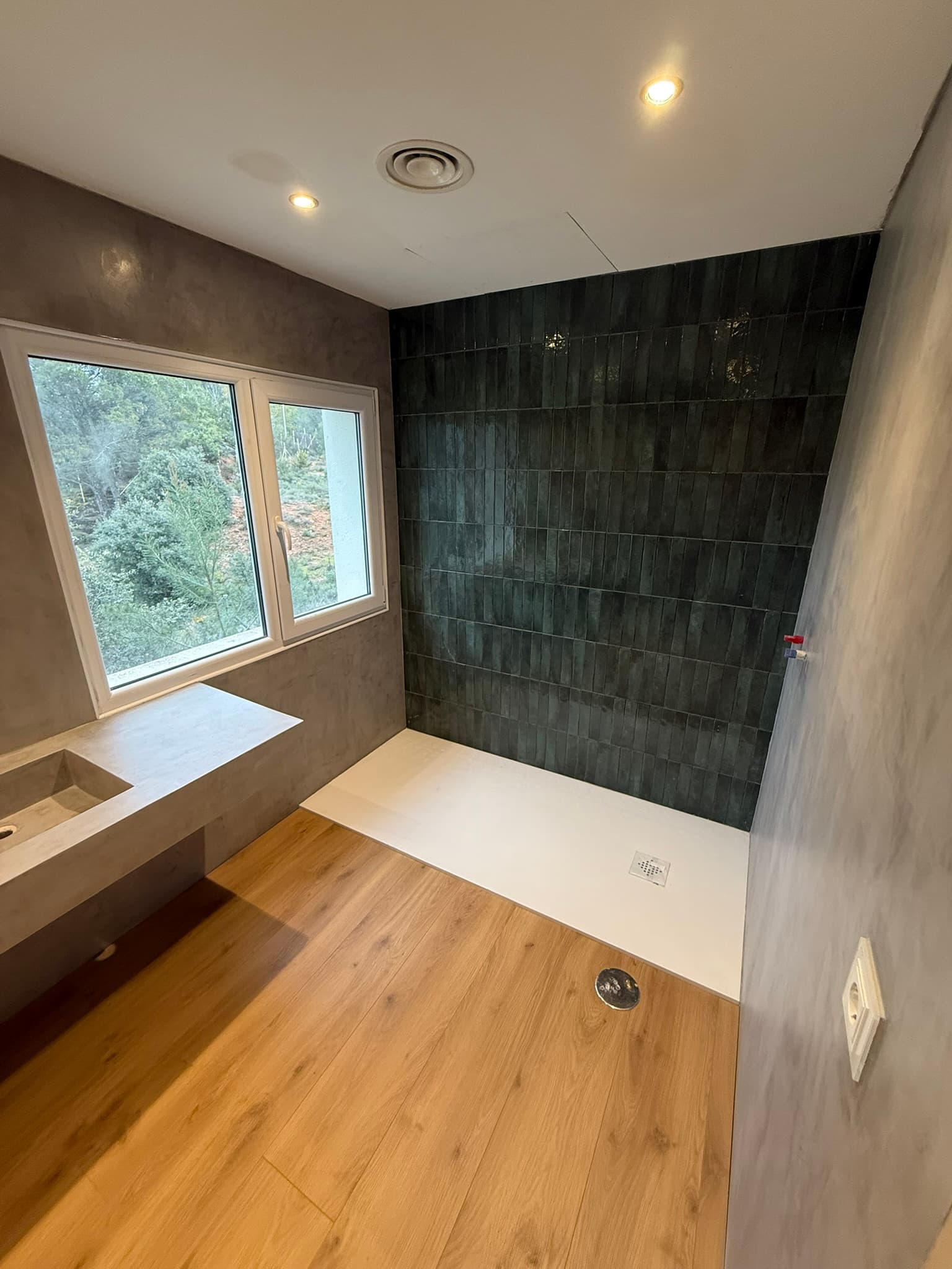 Ducha con pared de azulejo verde alargado combinada con microcemento gris en el resto del baño, junto a una ventana